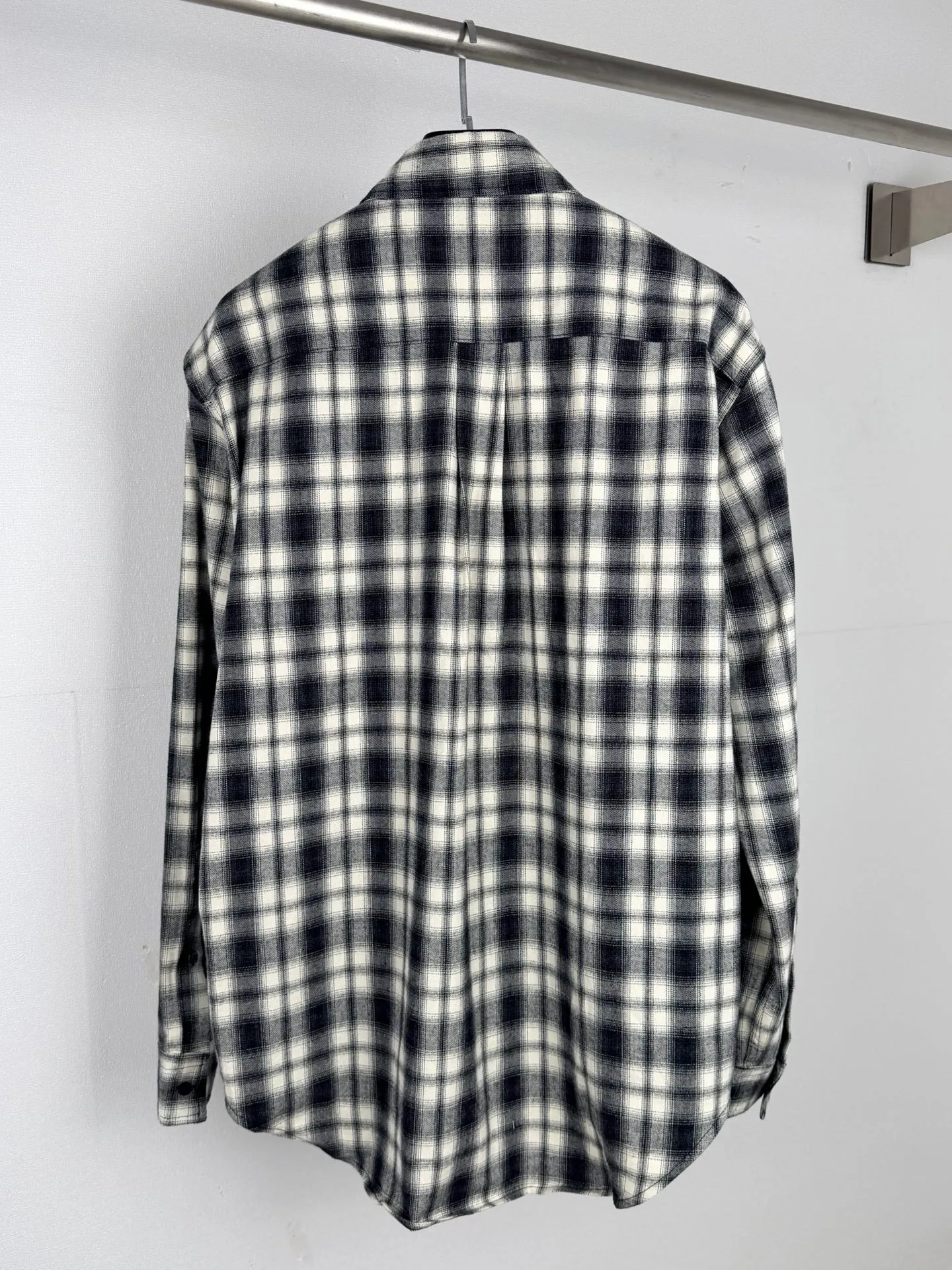 BALENCIAGA/balenciaga Black White plaid pocket embroidered logo shirt jacket The technical difficulties of this plaid shirt focus on fabric finishing, plaid matching and component sewing details. The fabric has been sanded and distressed to create a fine velvet feel on the surface, changing the original luster of the fabric and making the Black and White plaid colors transition softly. At the same time, the drape properties of the fabric itself are retained, taking into account the looseness of the upper body and the silhouette of the body, without appearing soft and shapeless. The biggest test of plaid garments comes from the alignment of the pieces. The front and rear body, large and small sleeves, and chest pockets are all independent pieces. In the cutting stage, the horizontal and vertical axes of the plaid need to be aligned, and the external chest bag is cut separately. The plaid on the bag surface needs to form a visual coordination with the main body of the garment. Cutting and typesetting will produce additional fabric loss, and there are high requirements for the cutting accuracy of the cutting bed typesetting and workers. The collar adopts a double-layer combined structure, and the collar surface and collar seat are double-stitched with even and regular stitching spacing to ensure the lapel's long-lasting shape and resistance to warping and deformation after repeated bending. The placket is sewn with regular topstitching and paired with Black matte buttons. The button points need to be aligned with the checkered lines to avoid damaging the overall visual order. The splicing of the cuffs also takes into account the grain direction, and the overlocking process reinforces the seams to improve the durability of the garment. After the entire garment is sewn, it also undergoes a garment washing process to further strengthen the Retro texture of the fabric, unify the tone and texture of the entire garment, weaken the harshness of industrial production, and finally present a relaxed Retro garment effect. Size: 46 48 50 52